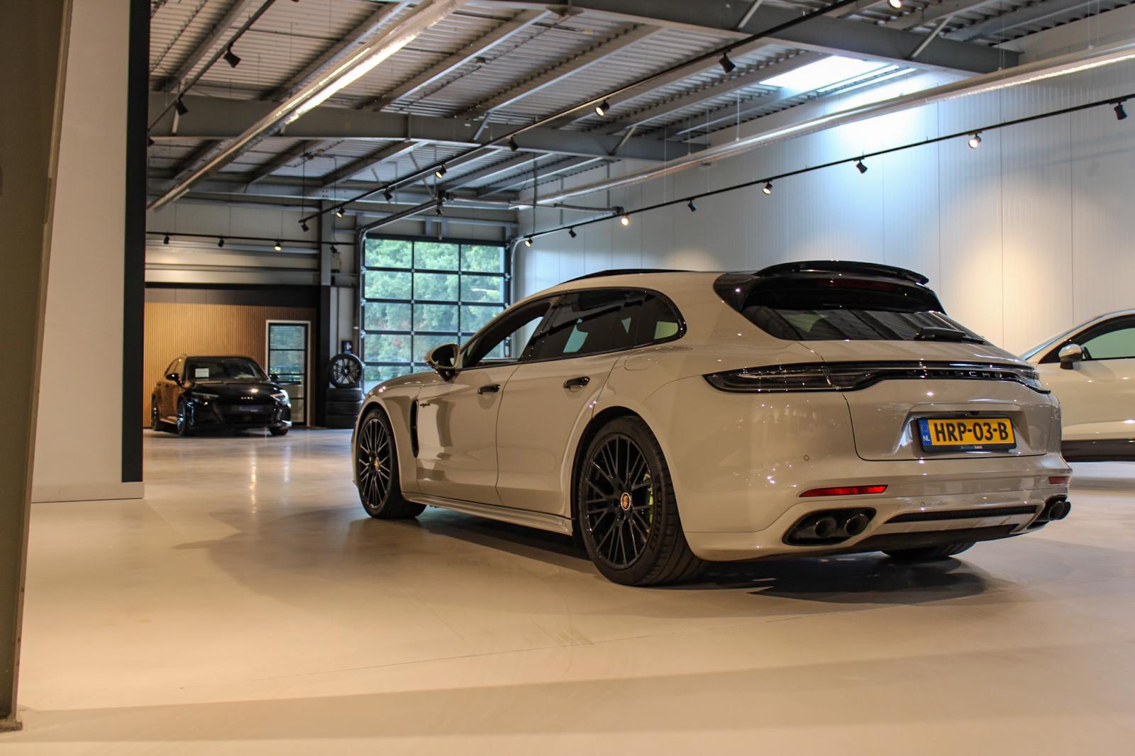 White Porsche station wagon parked inside a modern showroom with black alloy wheels and a visible license plate HRP-03-B.