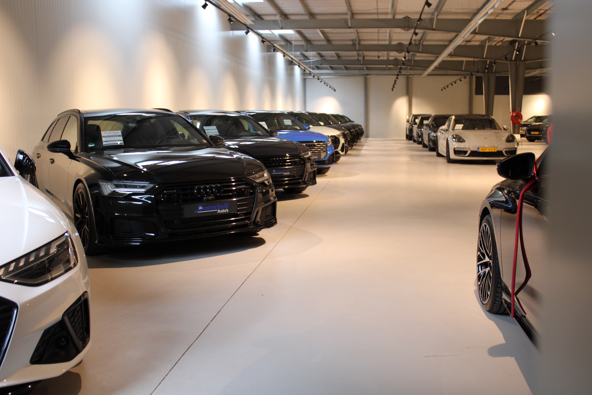 Indoor showroom with multiple luxury cars, including black and blue Audi models and a white Porsche, arranged in two rows.