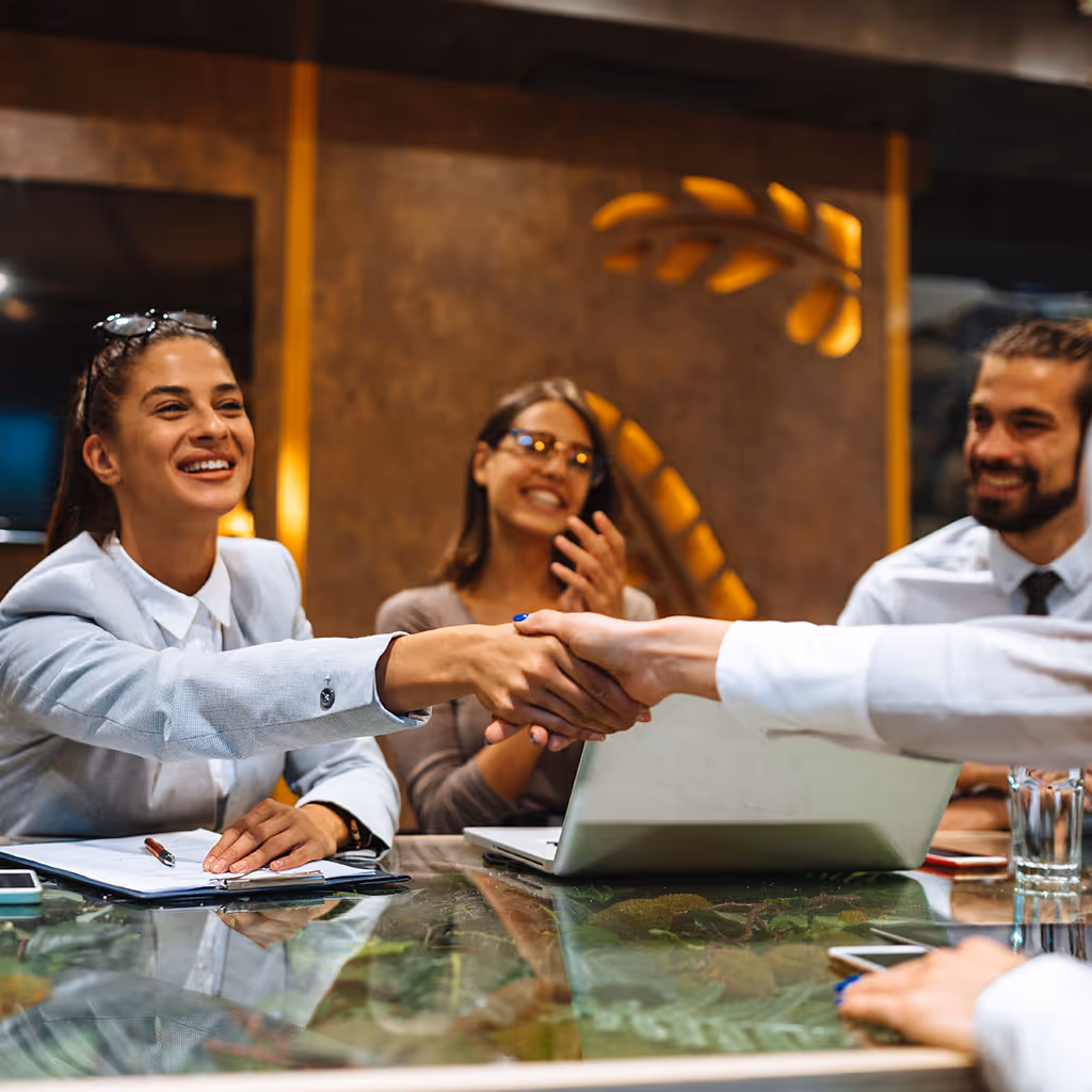 Two business professionals shaking hands over a glass table in a modern office with colleagues smiling nearby.