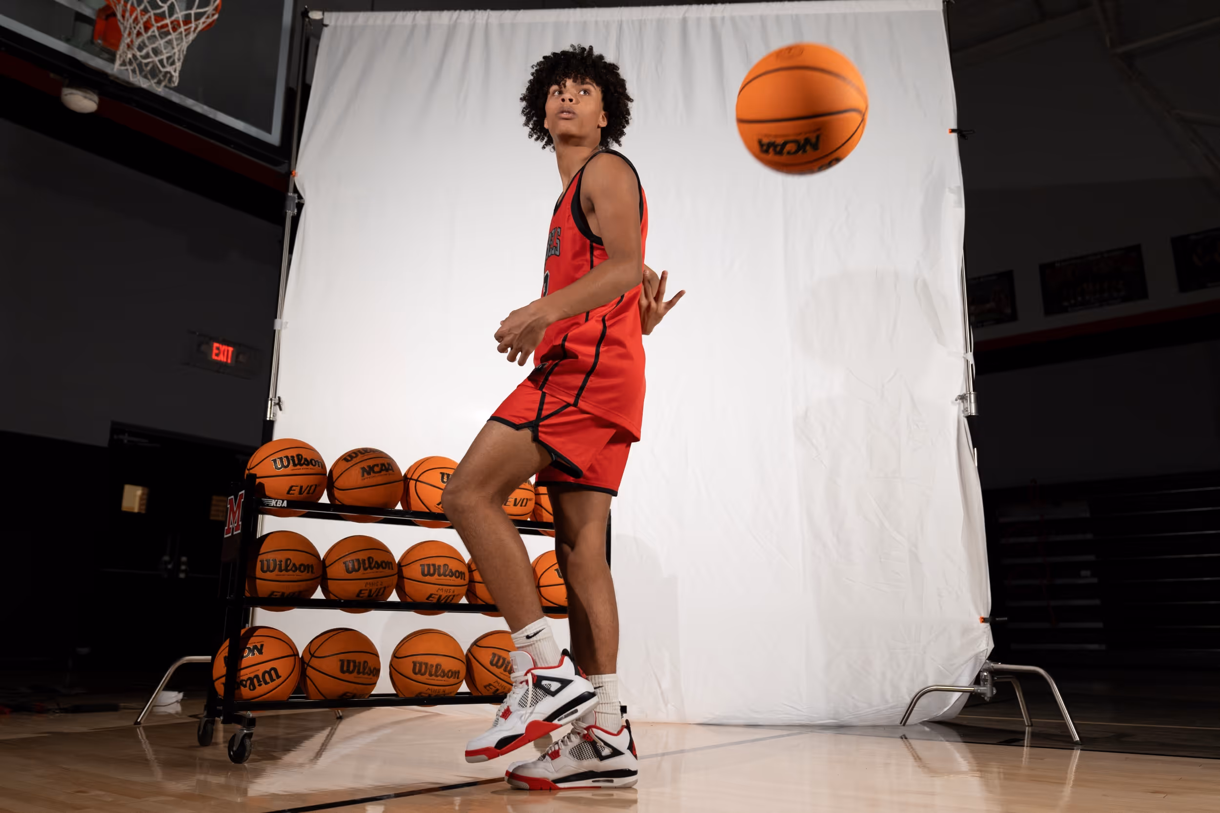 Young basketball player in red uniform and white sneakers interacts with a basketball in front of a white backdrop on an indoor court.