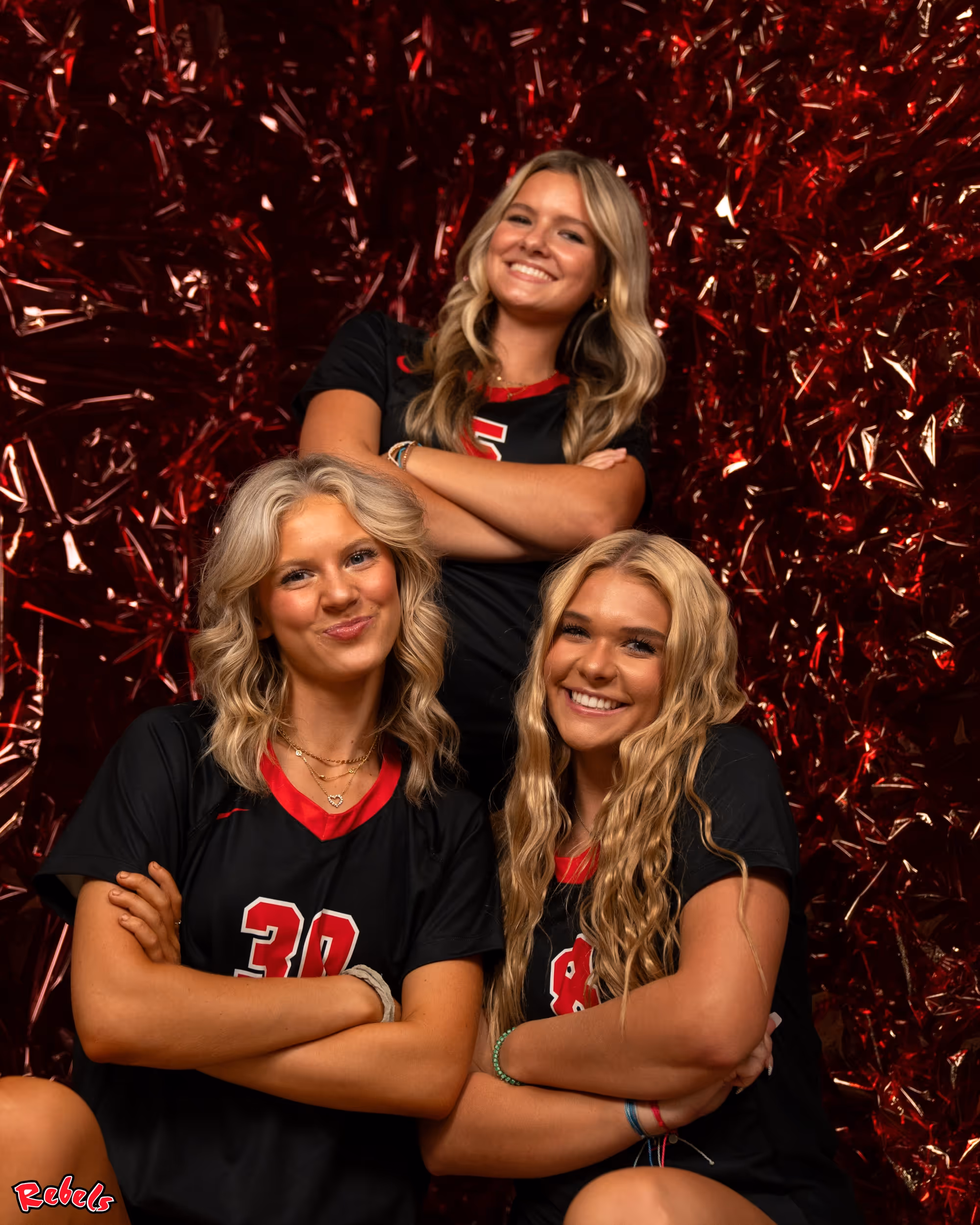 Three smiling female athletes in black and red jerseys posing with arms crossed in front of a red textured background.