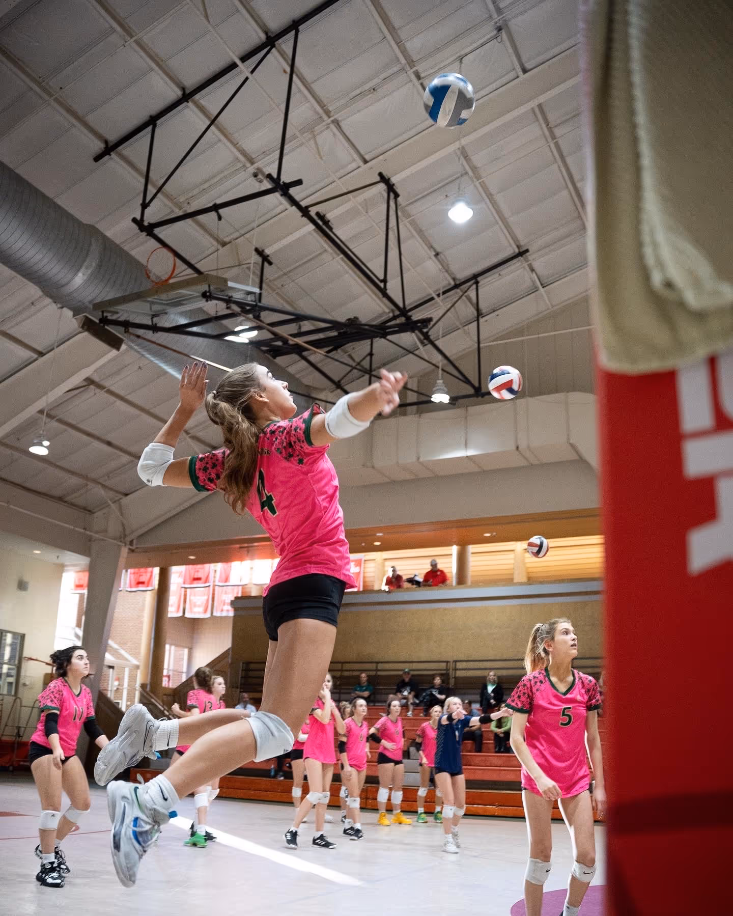 Female volleyball player in pink jersey jumping to hit the ball during an indoor game with teammates and spectators in the background.
