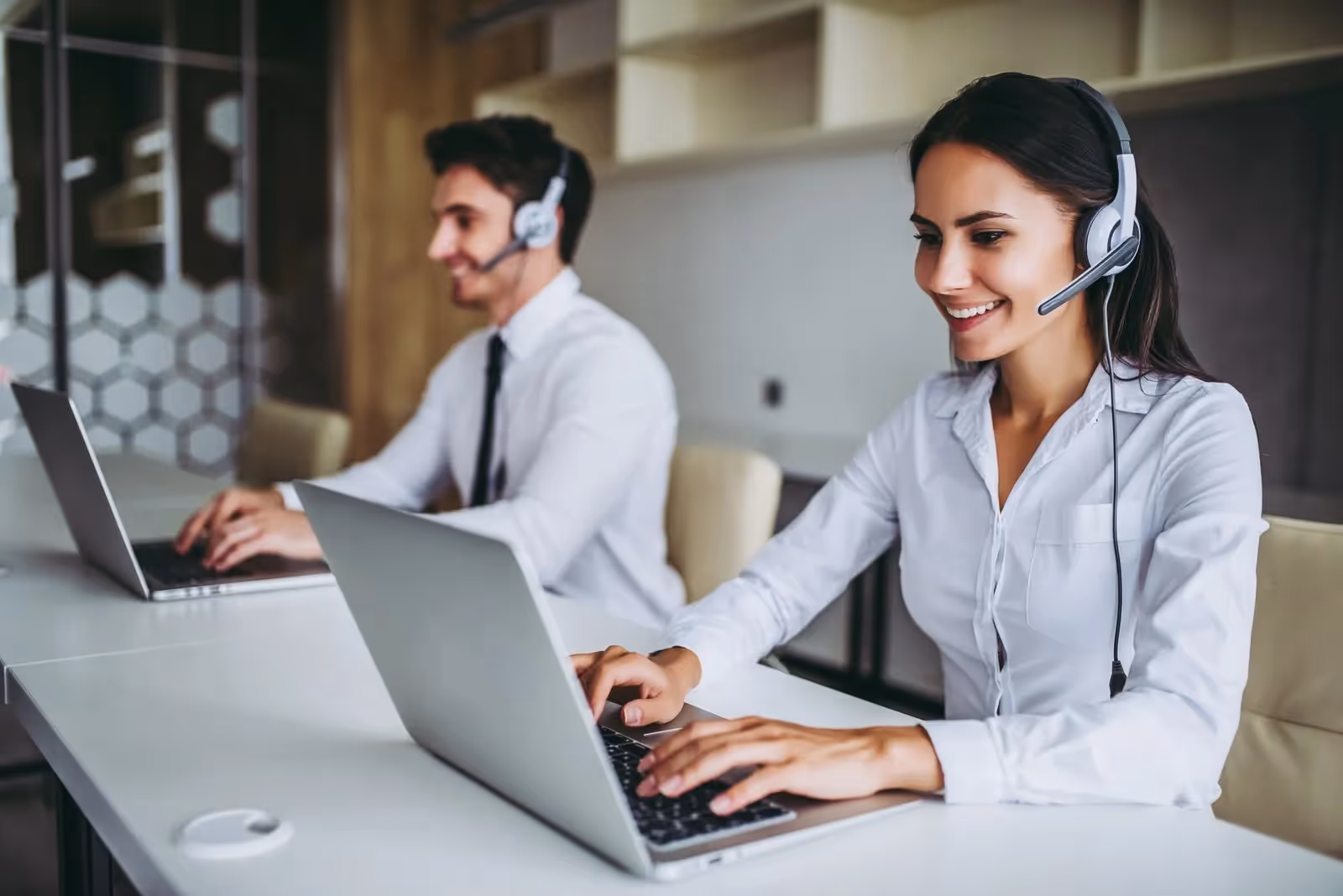 Dos agentes de centro de atención al cliente, un hombre y una mujer con auriculares, trabajando en portátiles y sonriendo.