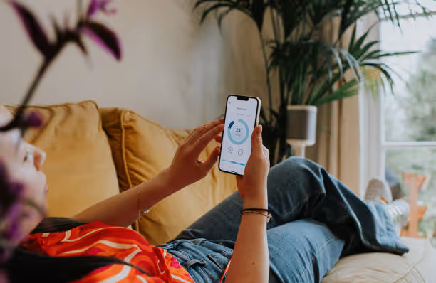 Persona recostada en un sofá amarillo usando un teléfono móvil para controlar la temperatura a 24 grados en una app.