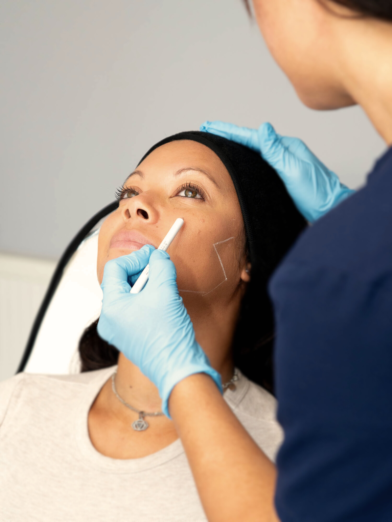 A woman with a black headband has white markings on her cheek while a person wearing blue gloves draws on her face with a white pencil.