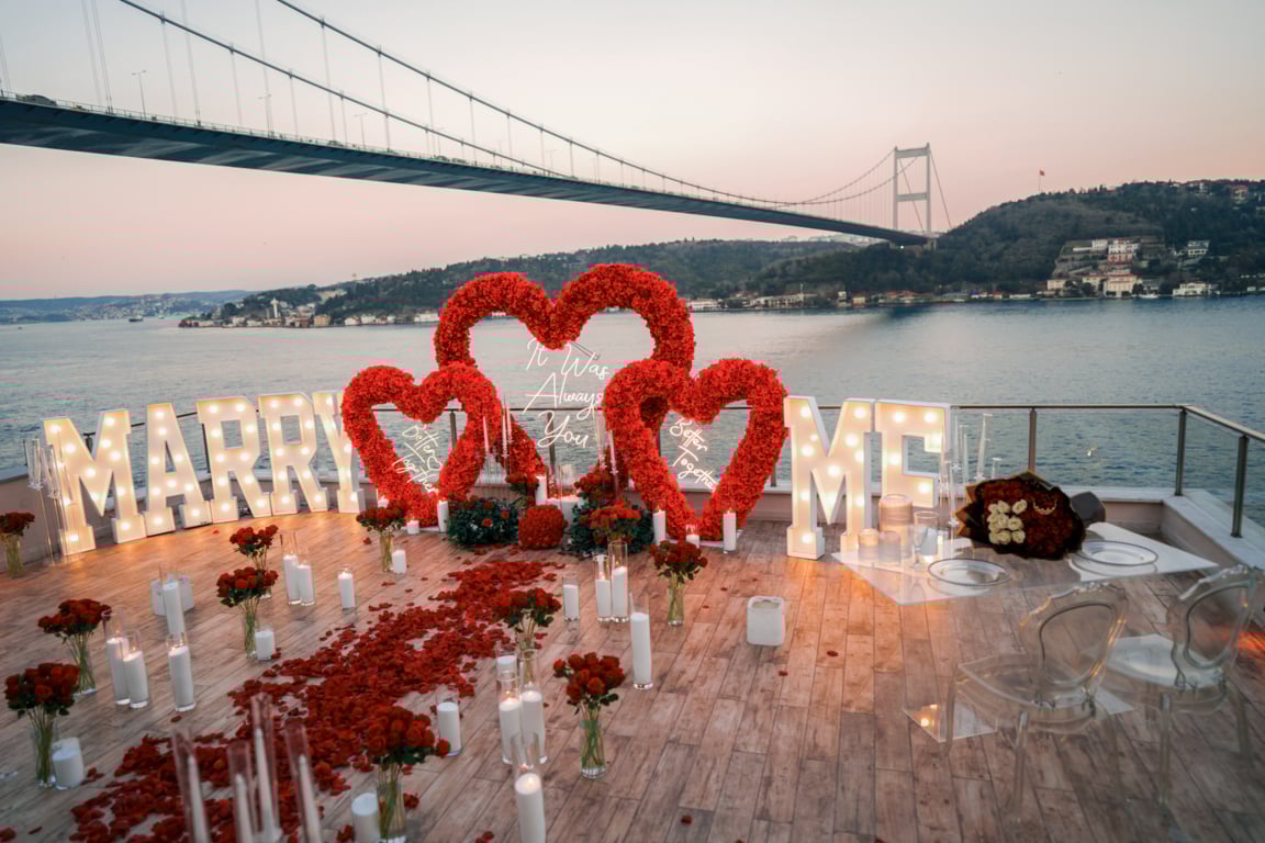 Romantic marriage proposal setup on a terrace by the water with illuminated 'Marry Me' letters, red heart-shaped floral arches, rose petals, and candles under a large bridge at sunset.