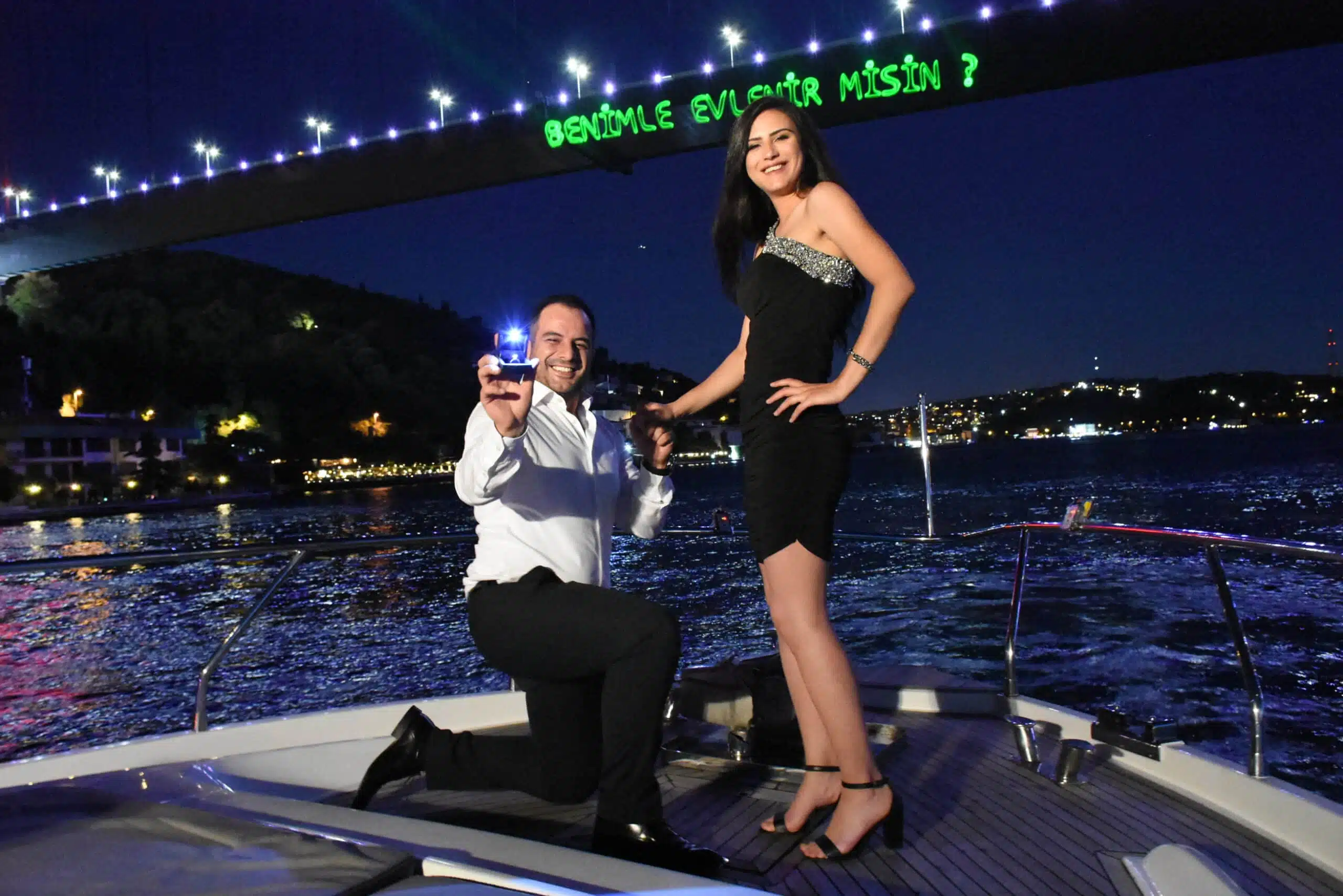 Man kneeling on a yacht at night, holding an open ring box proposing to a smiling woman standing in a black dress, under a lit bridge with green lights spelling a marriage proposal in Turkish.