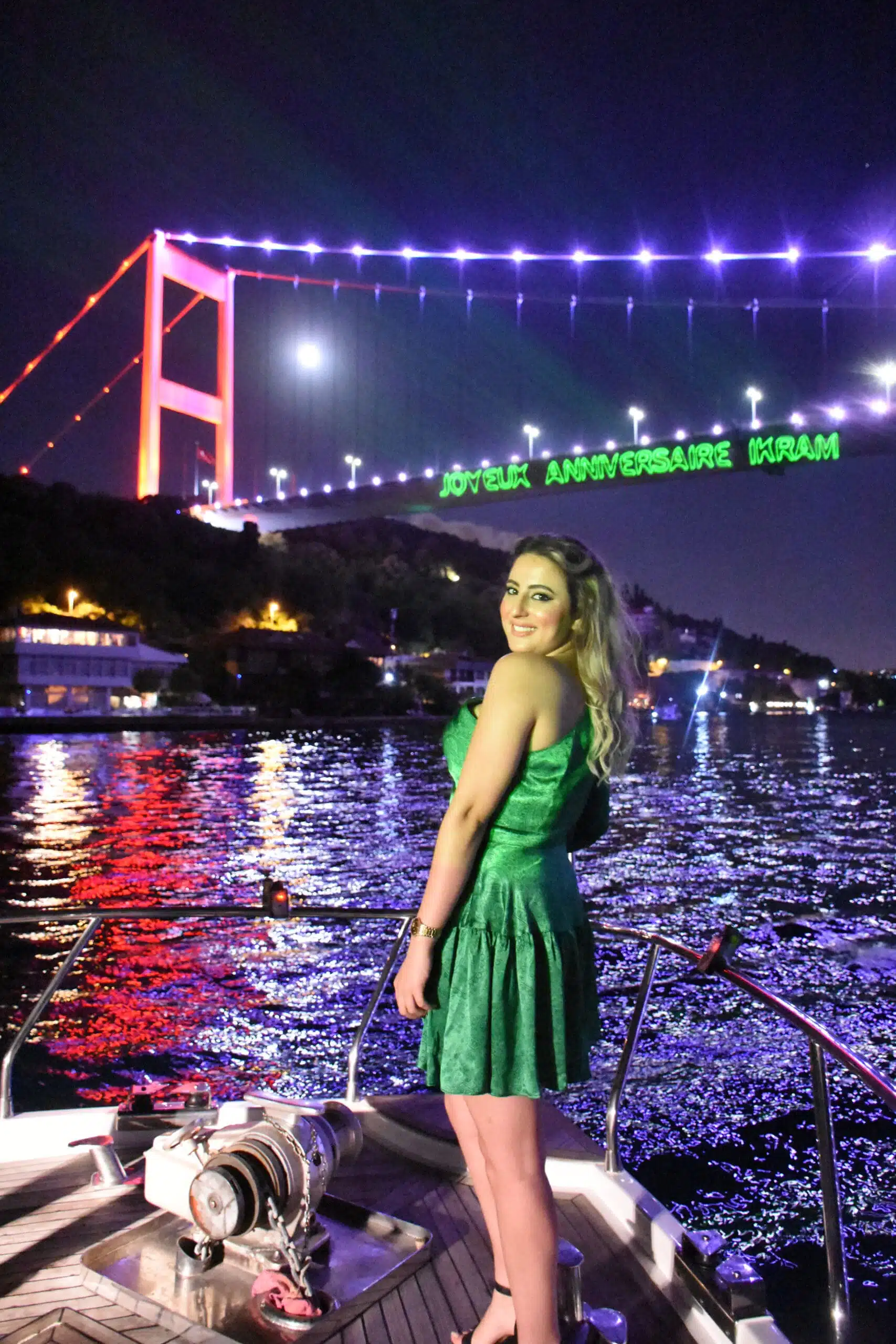Woman in a green dress standing on a boat at night with a lit suspension bridge and its colorful reflections in the water behind her.