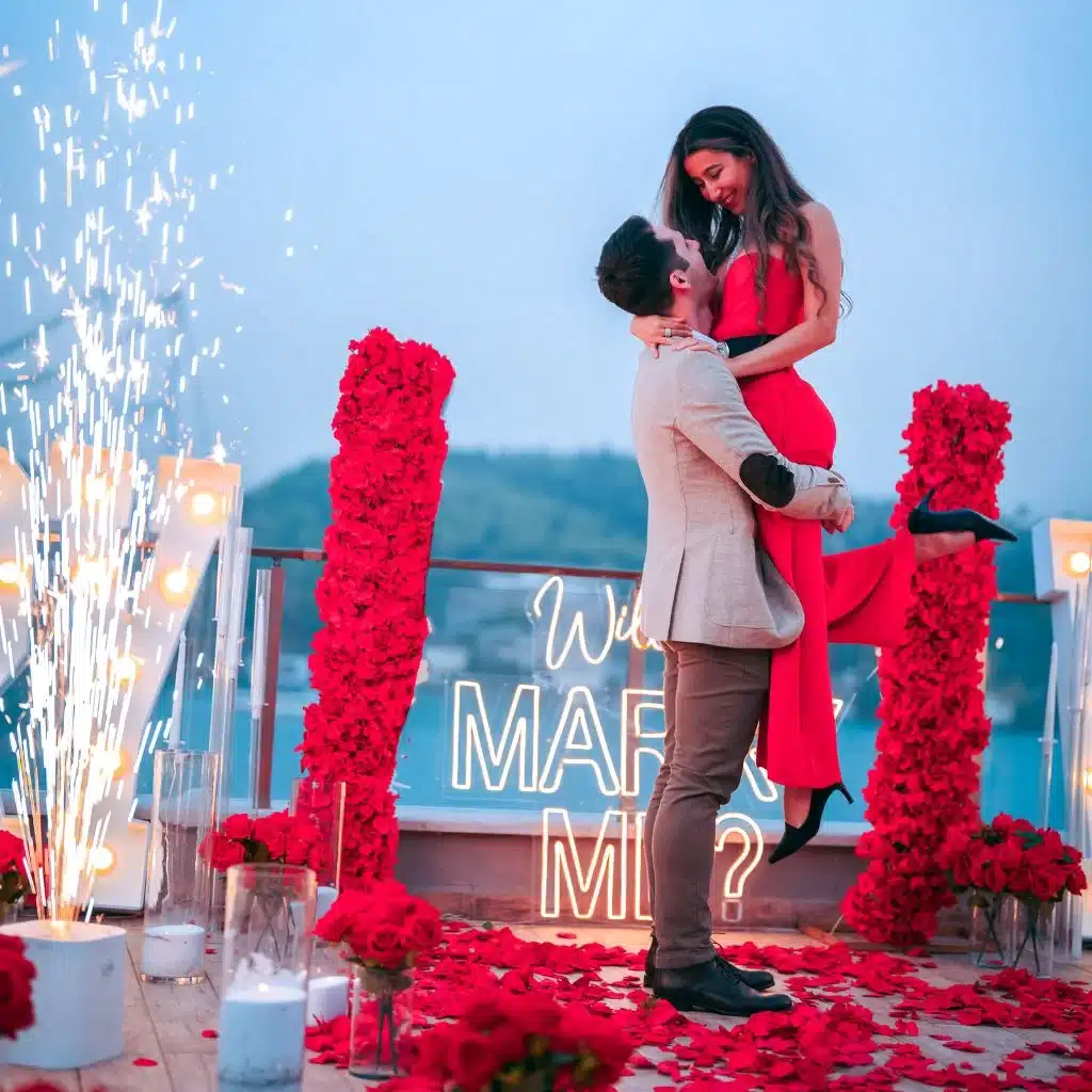 Man lifting woman in red dress surrounded by rose petals and fireworks with illuminated 'Will you marry me?' sign.