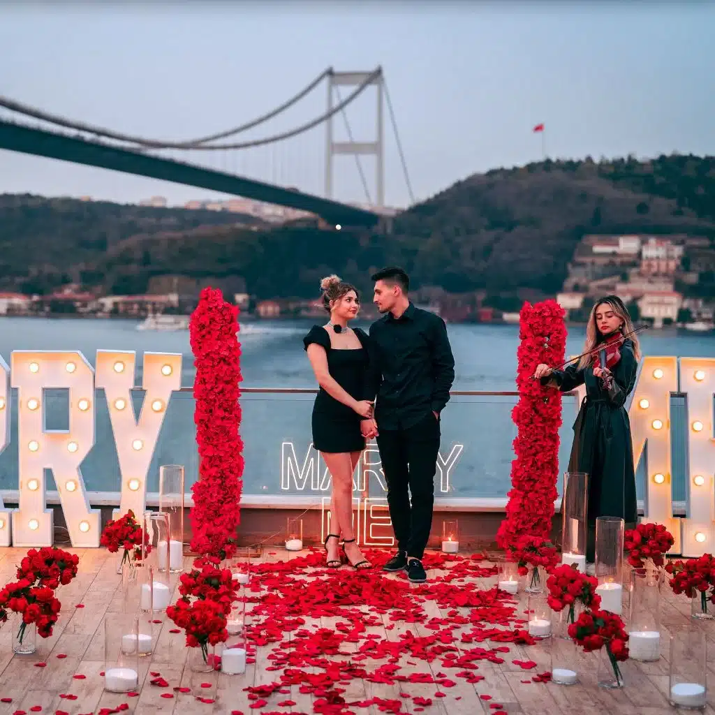 Couple standing on a rose-petal-covered deck with a violinist playing beside them and a bridge over water in the background.