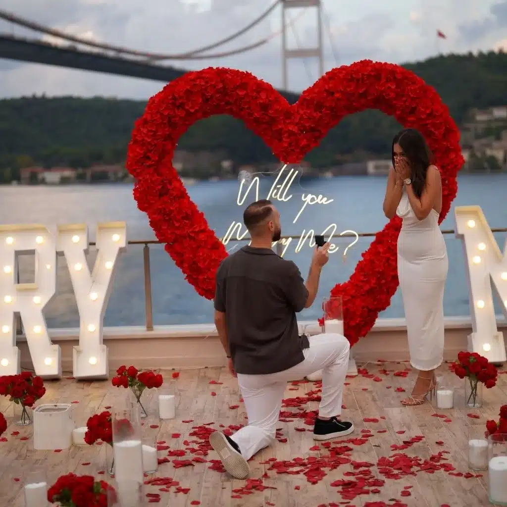Man kneeling with ring box proposing to surprised woman in white dress with large red rose heart and illuminated 'Marry Me?' sign by waterfront.