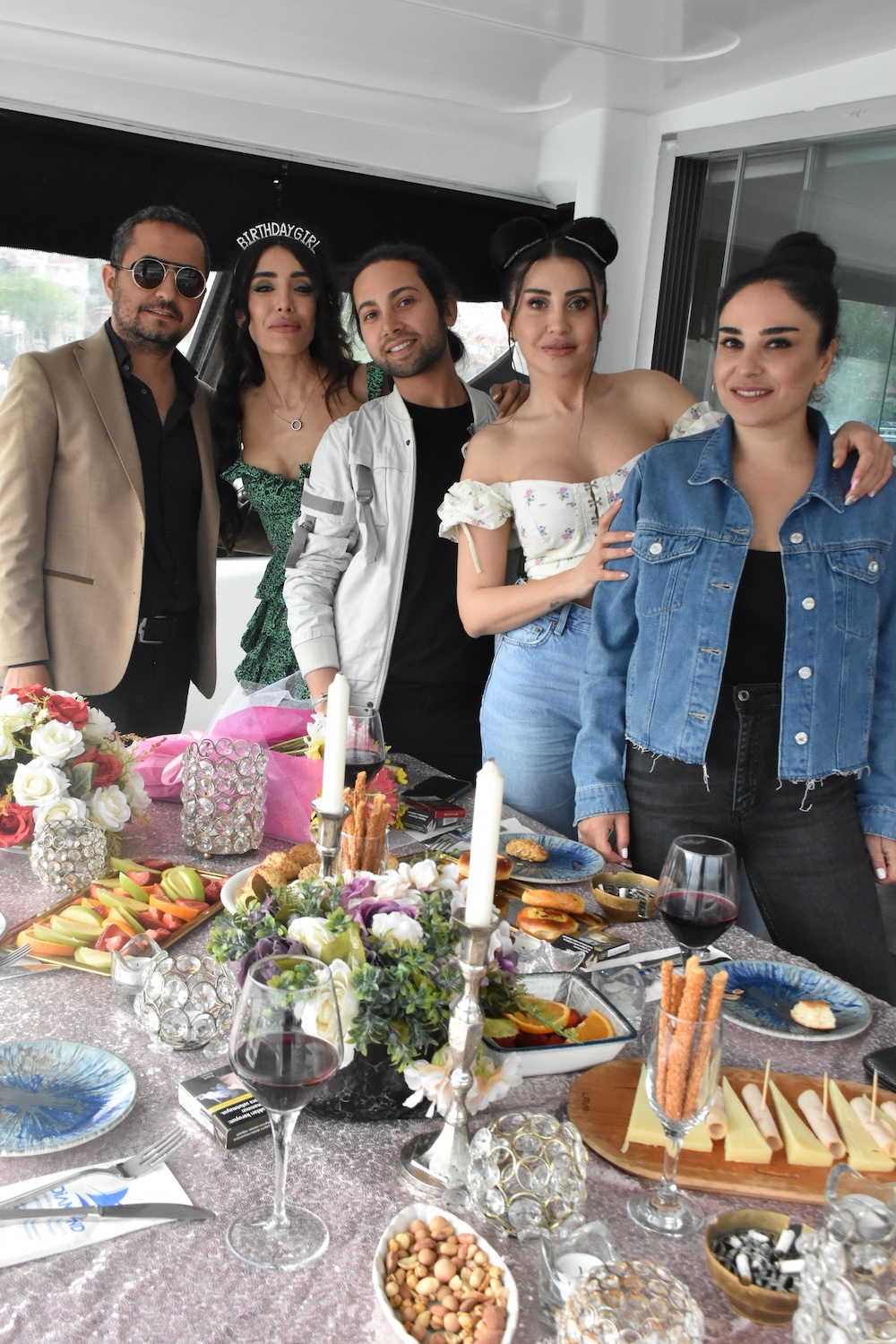 Five people posing closely together at a decorated birthday party table with flowers, candles, wine glasses, and assorted snacks.