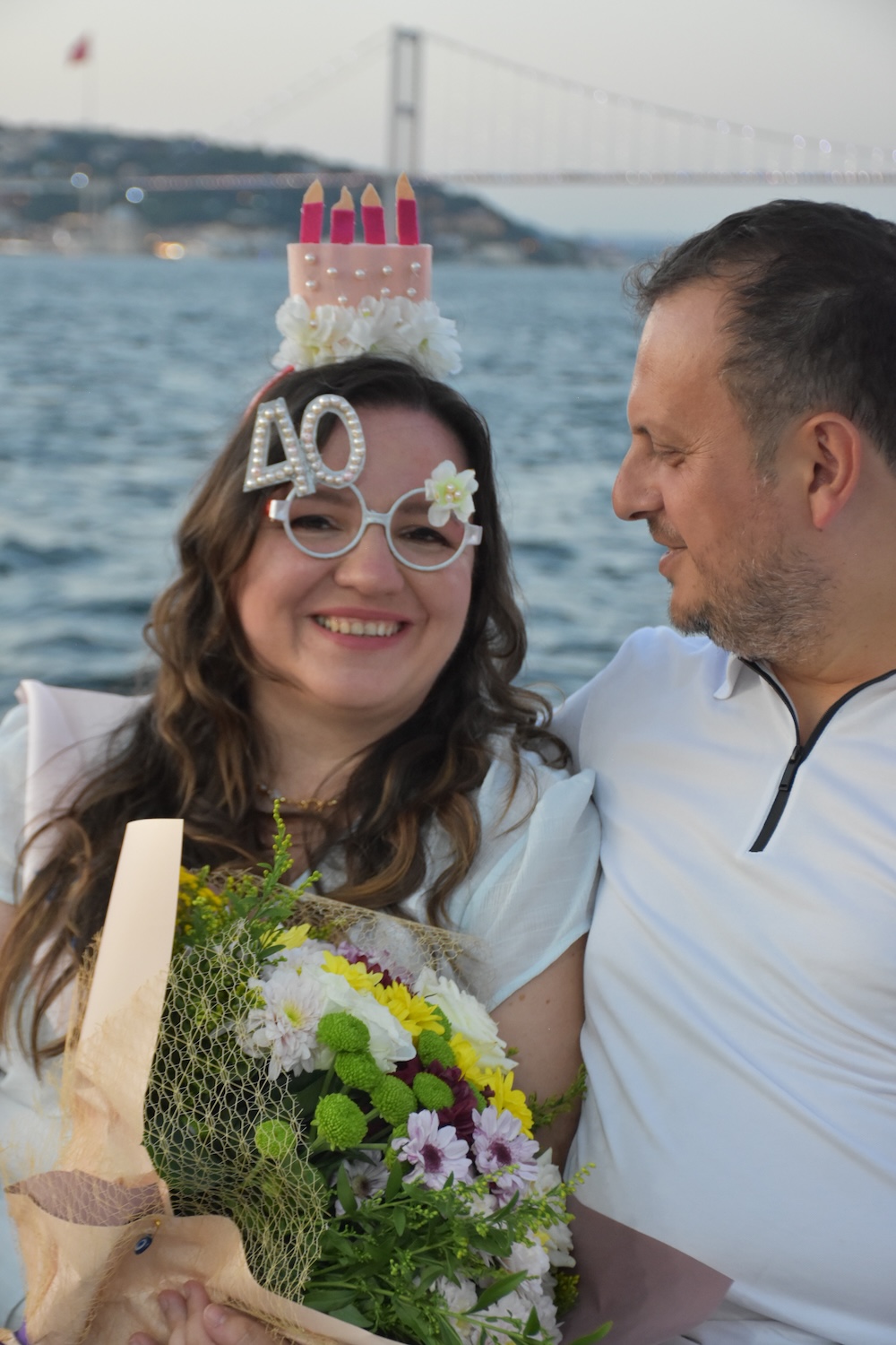 Smiling woman wearing novelty 40th birthday glasses and a cake headband holding a bouquet, with a man looking at her by the water with a bridge in the background.