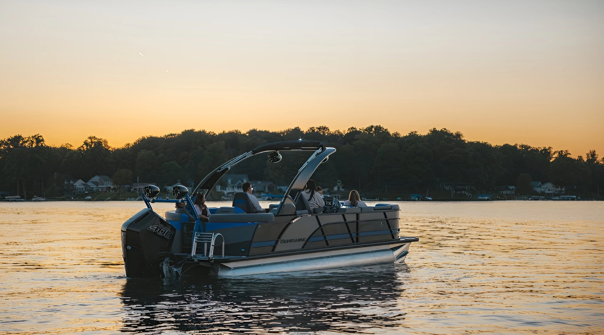 Pontoon boat with people cruising on calm lake water at sunset with tree-lined shore in the background.