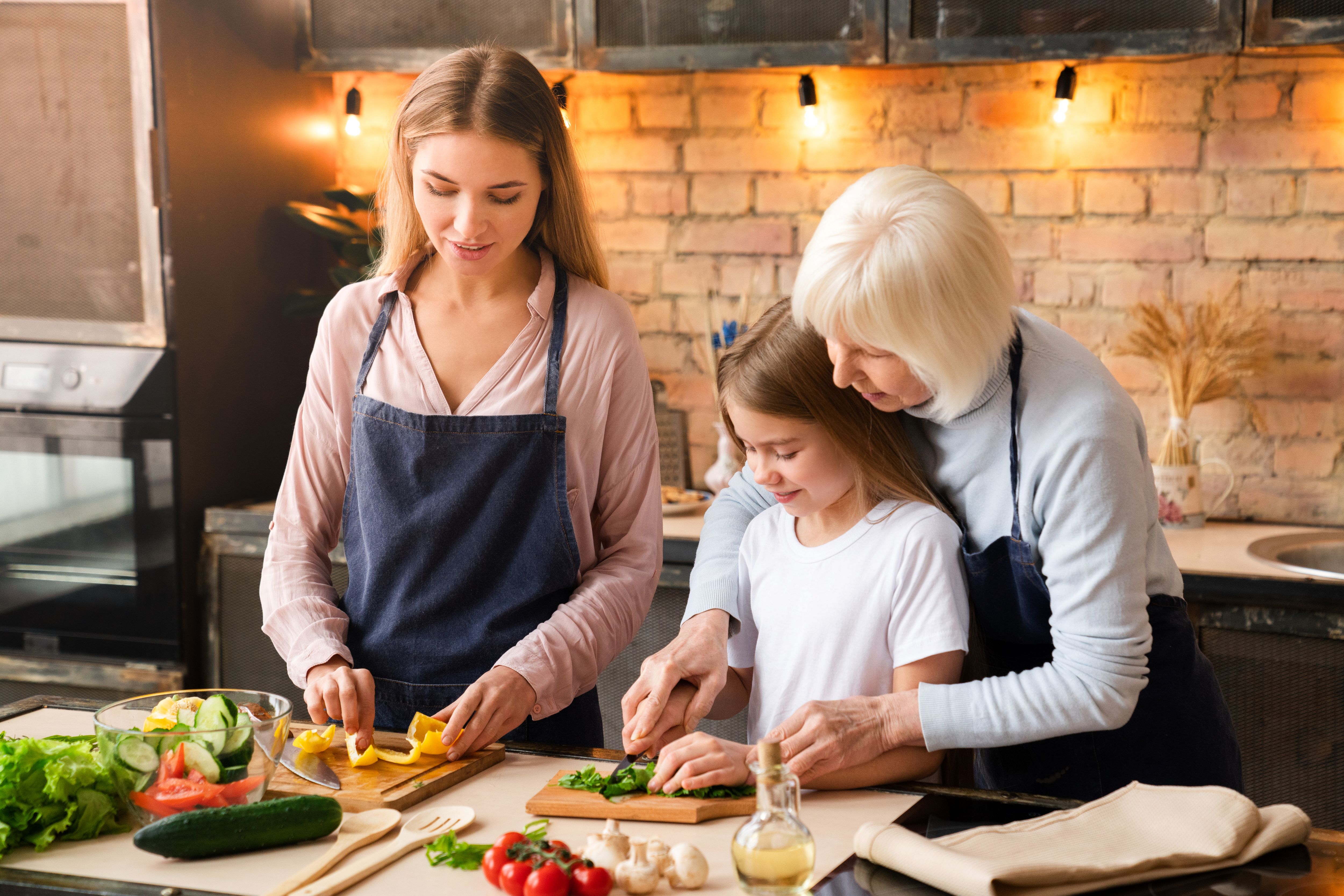 Drei Generationen – Großmutter, Mutter und Tochter – schneiden gemeinsam frisches Gemüse in einer warmen, hellen Küche. Das Bild vermittelt Familiennähe, gesunde Ernährung und gemeinsames Aktivsein.