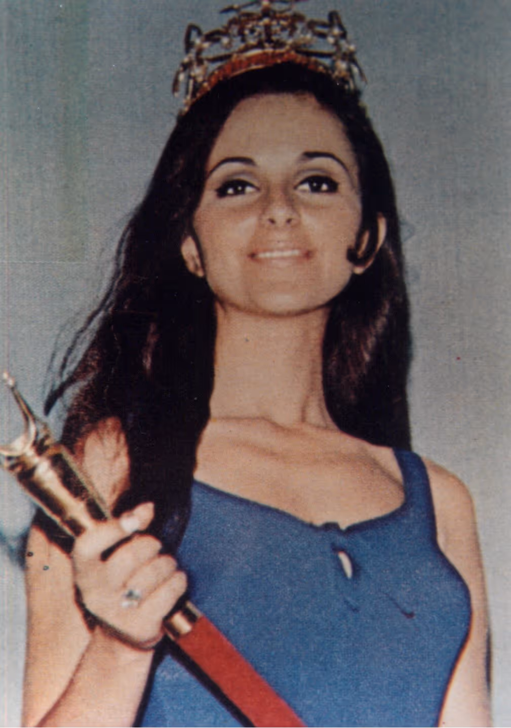 Mujer joven con corona y vestido azul sosteniendo un cetro dorado con una sonrisa.