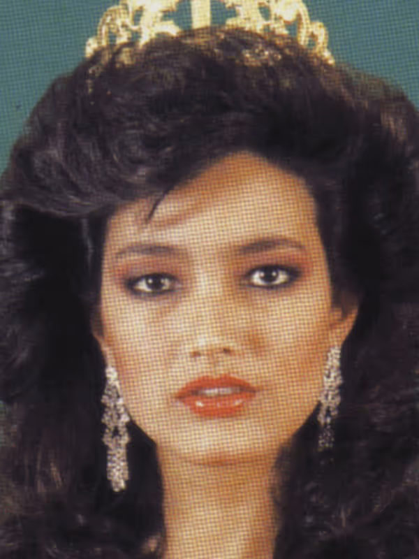 Mujer con corona dorada, cabello rizado oscuro, labios rojos y aretes largos plateados.