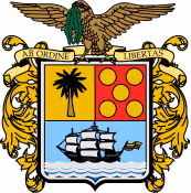 Escudo de la Gobernación de Bolívar con un águila dorada, un escudo dividido en cuatro partes con un árbol, un barco, y círculos rojos, y la frase 'Ab Ordine Libertas'.