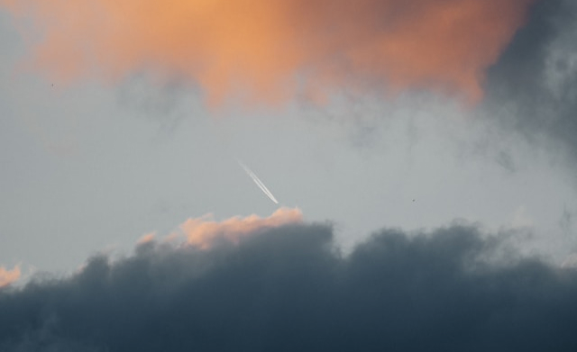 Sky with dark clouds at the bottom and soft orange-tinted clouds at the top, with a faint white airplane contrail in the center.