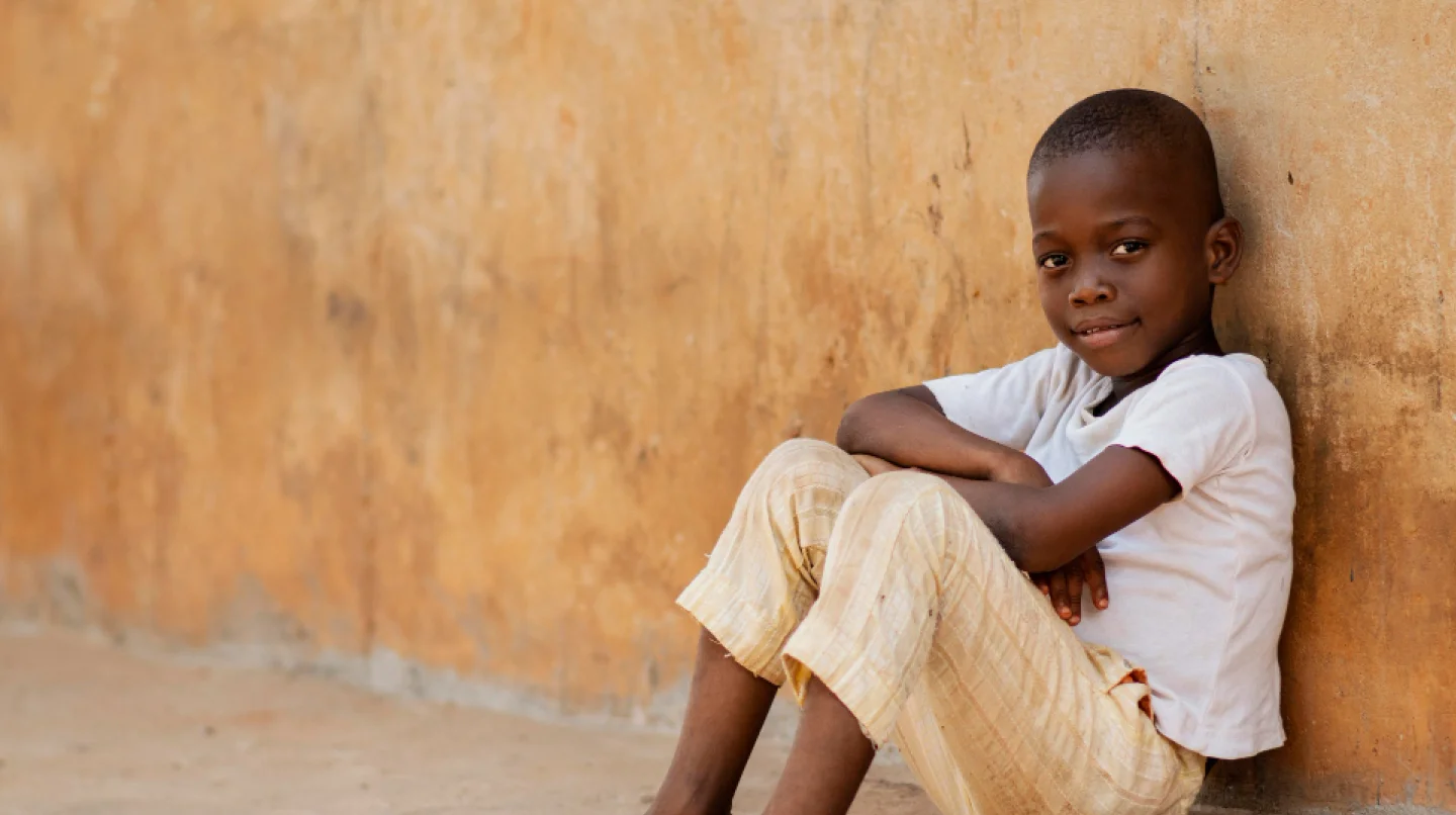 Young boy sitting on the ground with arms crossed, leaning against a textured orange wall, smiling softly.