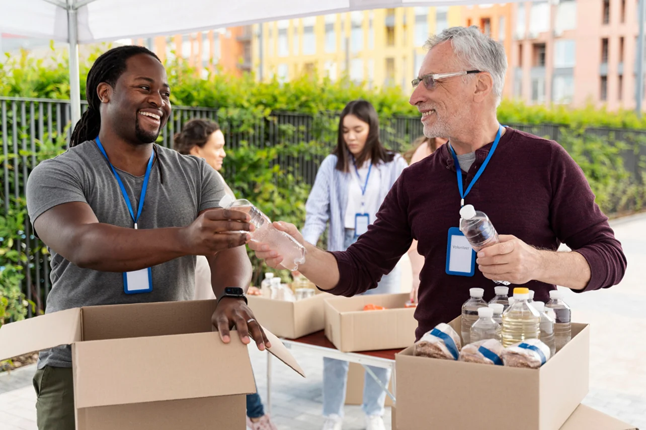 Two male volunteers exchanging a bottle of water while packing boxes with supplies outdoors under a tent.