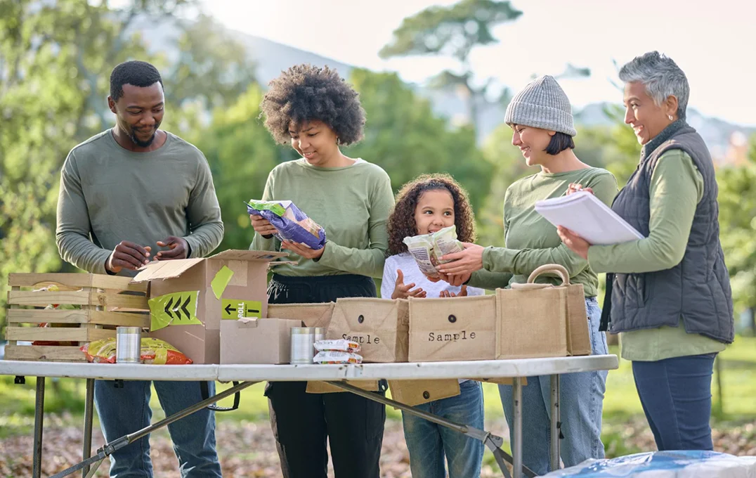 Five diverse volunteers outdoors sorting and packing food items on a table with boxes and reusable bags.