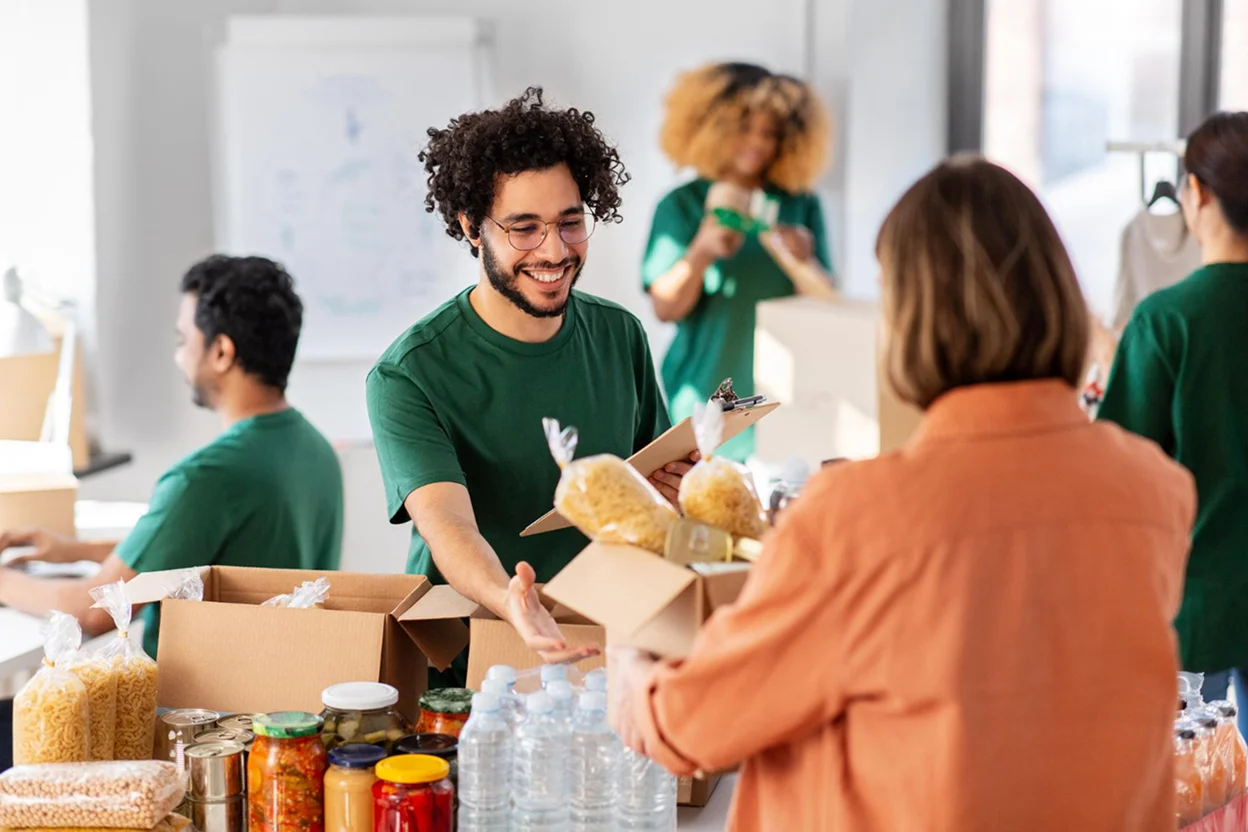 Volunteers packing food items like pasta, bottled water, and canned goods into boxes in a bright room.
