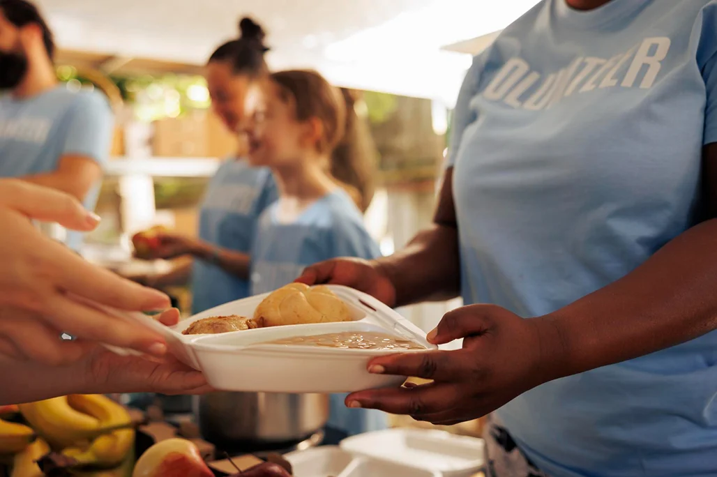 Volunteer in a blue shirt handing a food tray with bread roll and meal to a person at a community food service.