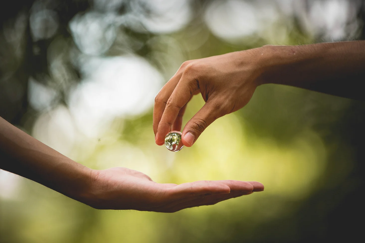 Hand holding a small clear glass ball above an open palm with blurred green background.