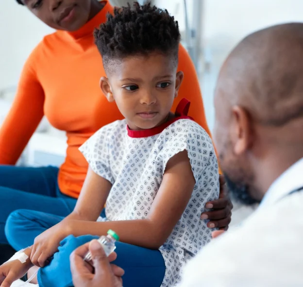 A young boy in a hospital gown sitting with a woman in an orange top while a doctor offers him a vial of medication.