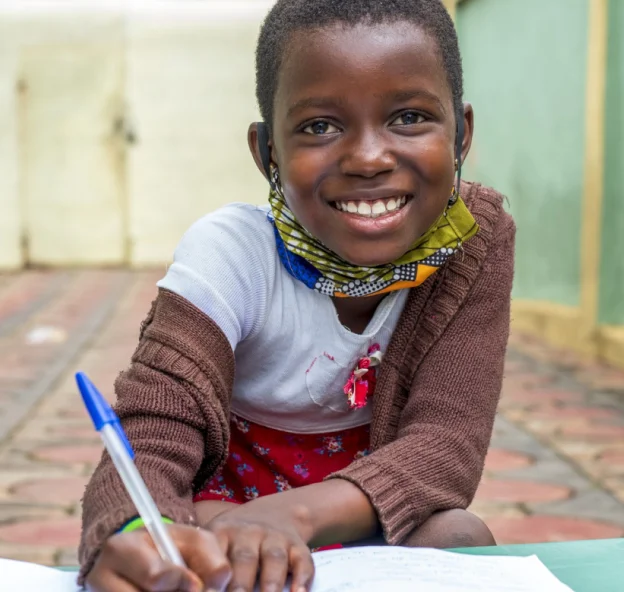 Smiling young girl writing with a blue pen while sitting on a tiled floor, wearing a brown sweater and a colorful mask under her chin.
