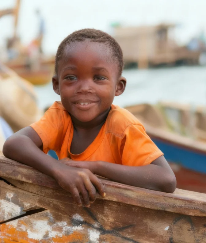 Smiling young boy wearing an orange shirt leaning on the edge of a wooden boat near a body of water.