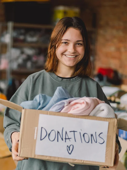 Smiling young woman holding a cardboard box labeled 'DONATIONS' filled with folded clothes.