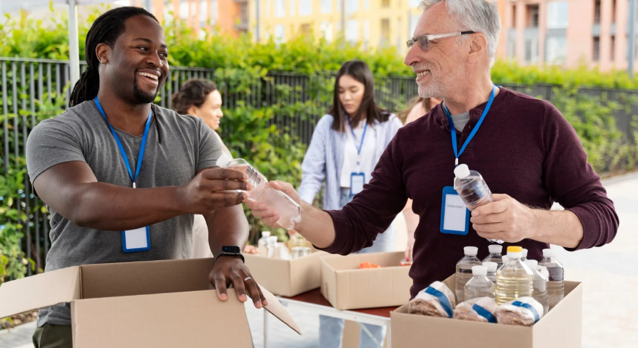 Two volunteers smiling and handing out bottled water from cardboard boxes at an outdoor community service event.