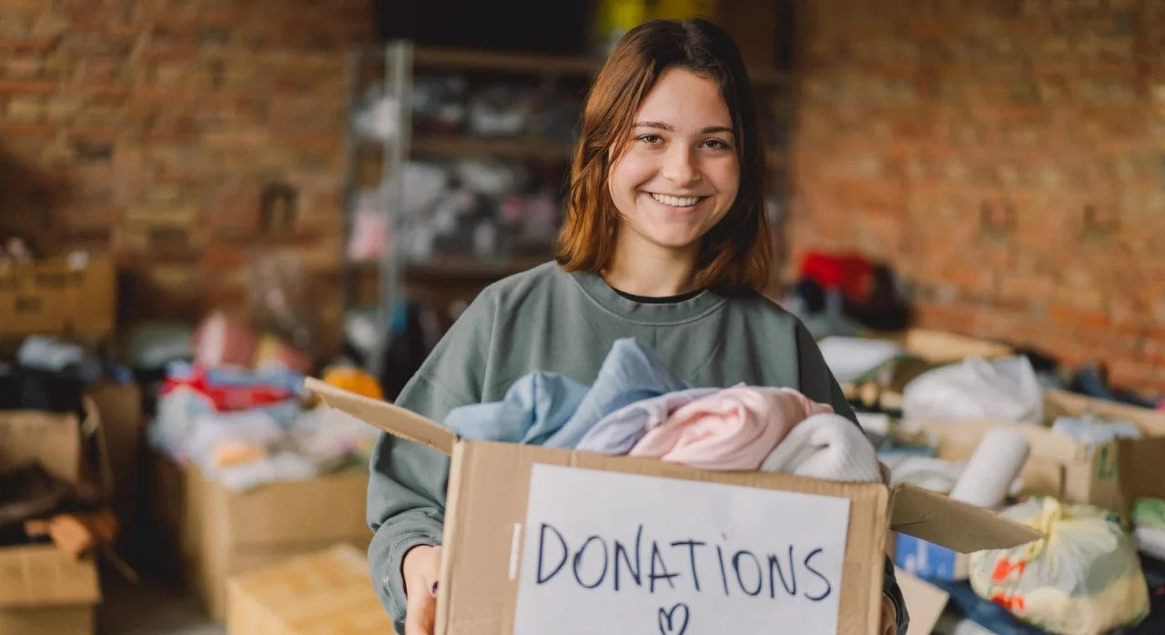 Smiling young woman holding a cardboard box labeled donations filled with clothes in a room with other boxes and donations.