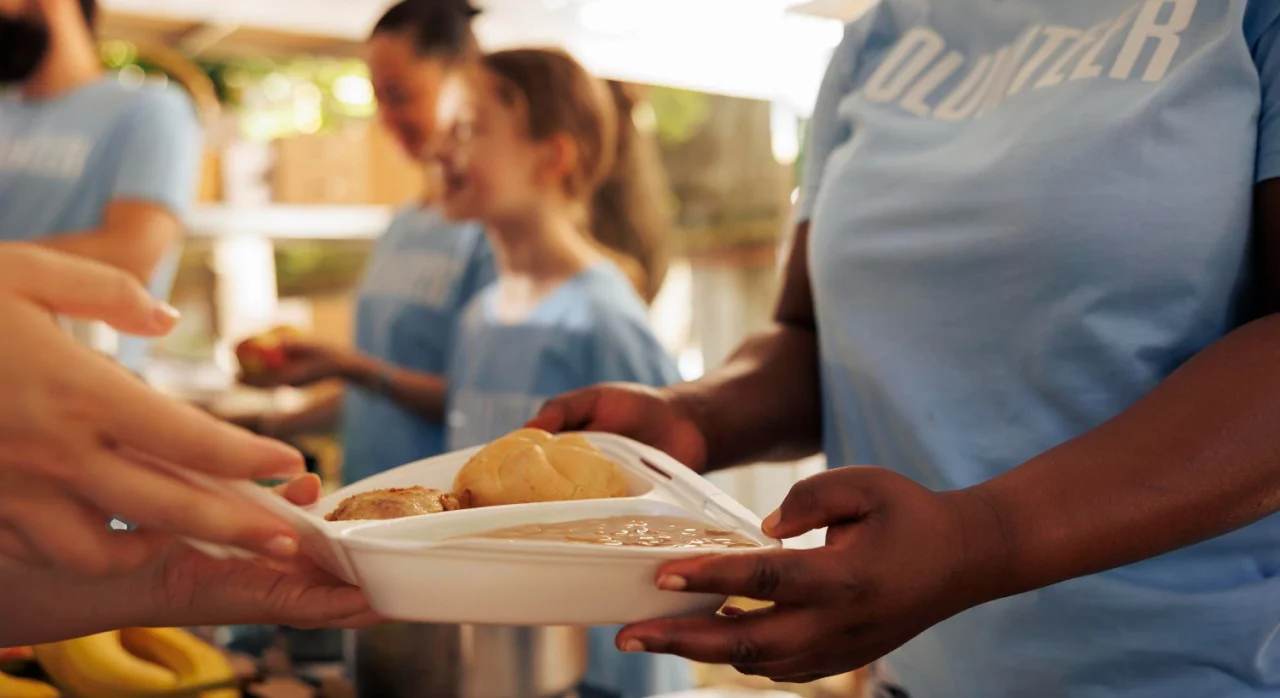 Volunteer handing a plate with food including biscuits and beans to another person at a community food service.