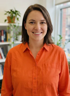 Smiling woman with shoulder-length brown hair wearing an orange button-up shirt in a bright room with books and plants in the background.