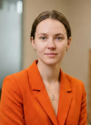 Portrait of a woman with brown hair wearing an orange blazer and a necklace with a pendant.