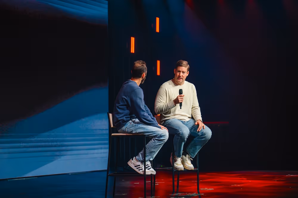Two men sitting on stools on stage, one holding a microphone during a conversation under stage lighting.