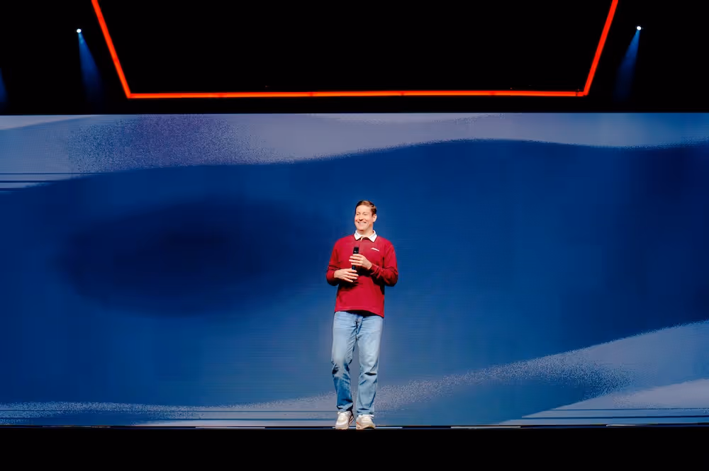 Rory Nicol in a red sweater and jeans speaking on stage with a microphone, standing before a large blue abstract background and a red-lit ceiling feature.