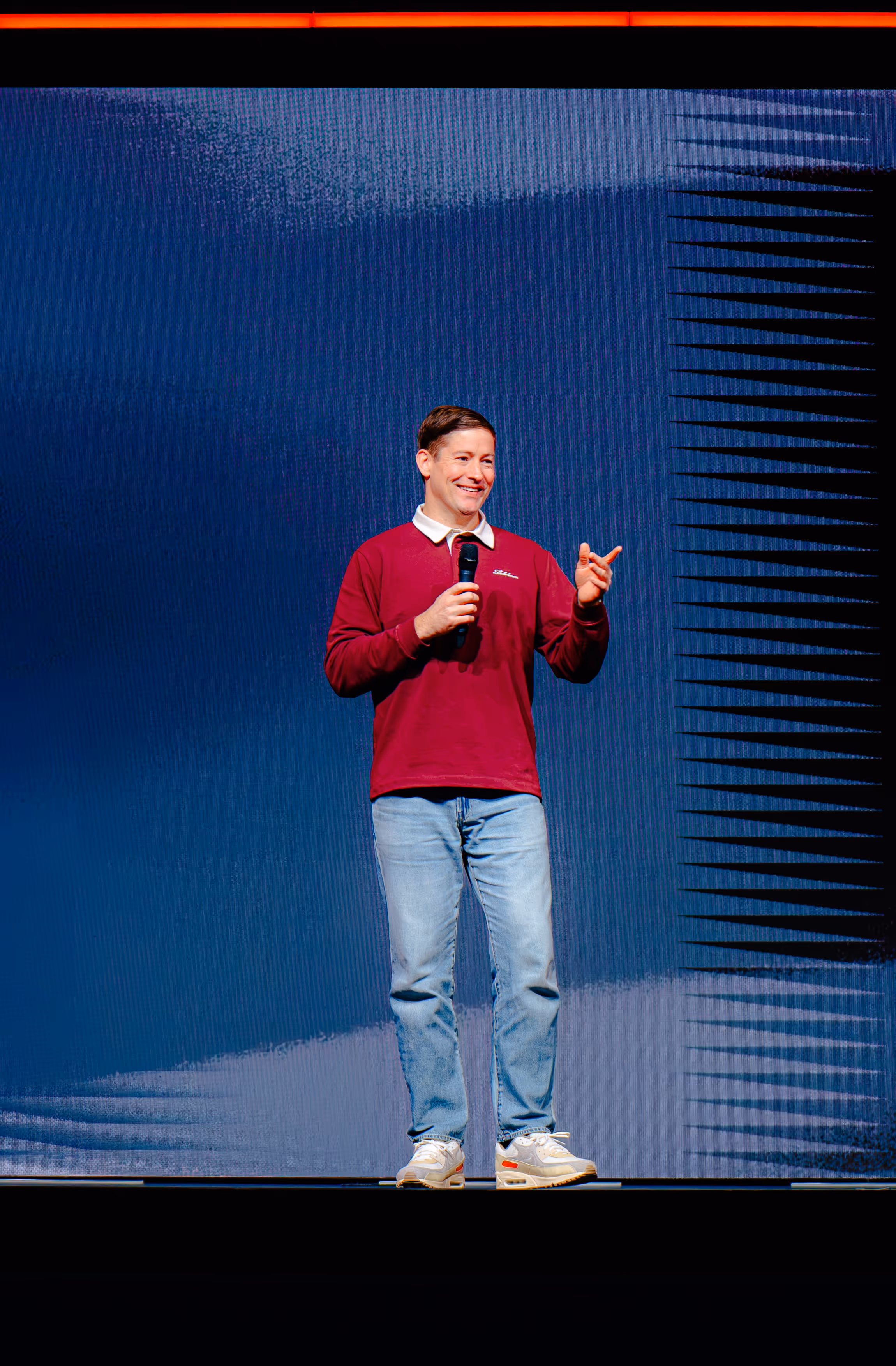 Rory Nicol in red sweatshirt and jeans speaking on stage holding a microphone with a blue and black abstract background.