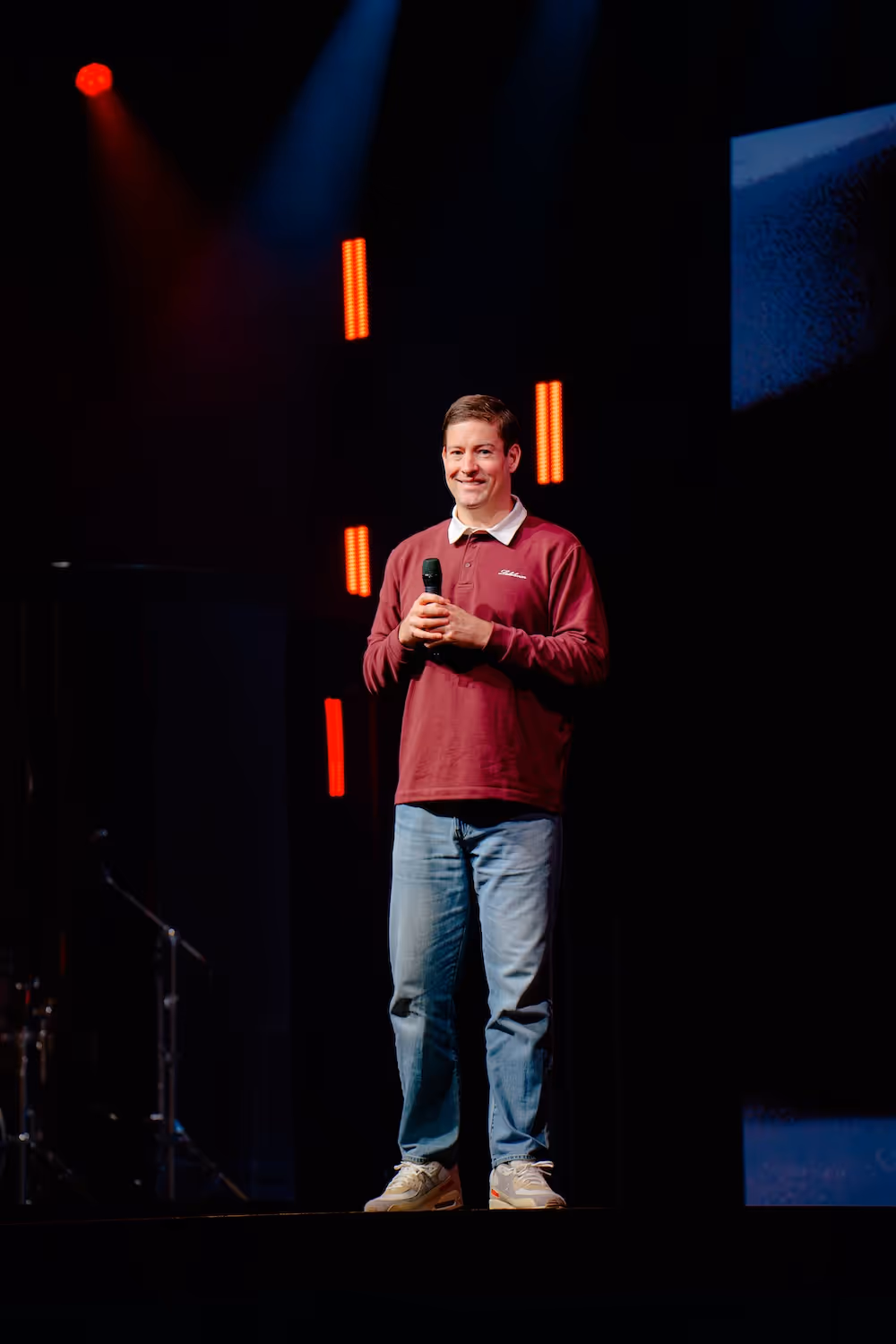 Rory Nicol wearing a maroon long-sleeve shirt and jeans holding a microphone on stage with dark background and red vertical lights.