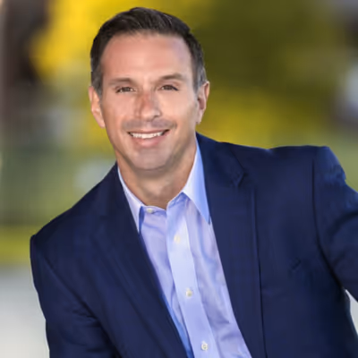 Damon West in a blue suit jacket and light blue shirt against a blurred outdoor background.