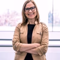 Elaine Bonner with glasses and shoulder-length hair wearing a tan jacket over a black top, standing with arms crossed indoors near a large window.