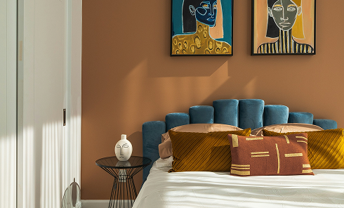 Modern bedroom with blue velvet upholstered headboard, white bedding, brown and mustard pillows, round side table with a white sculptural vase, and abstract face paintings on a brown wall.