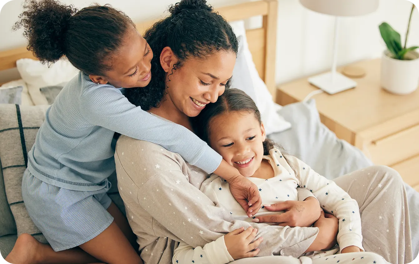 Mother sitting on bed hugging two smiling daughters in pajamas, one daughter hugging her from behind.