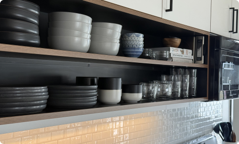 Open kitchen shelves with stacked black and white bowls, patterned cups, glasses, and cookbooks above a tiled backsplash.