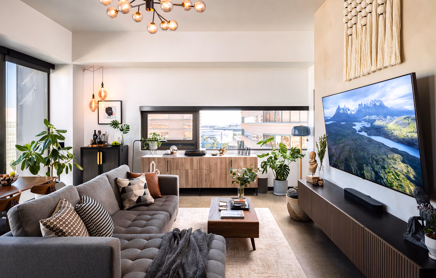 Modern living room with gray sofa, wooden furniture, plants, and a large TV showing a mountain landscape.