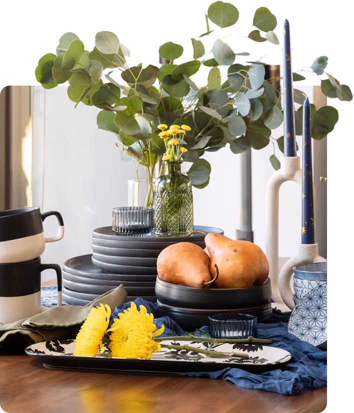Table setting with pears, yellow flowers, green leaves, candles, stacked plates, and cups on cloth.