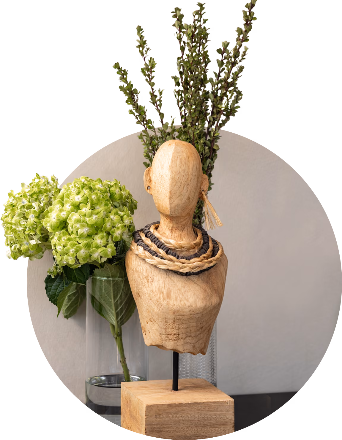 Wooden mannequin bust with braided necklaces in front of green hydrangea and leafy plants in vases.